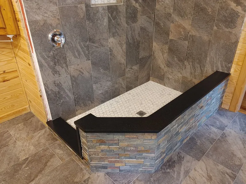 Angled shower curb wall capped in black stone over stacked stone tile