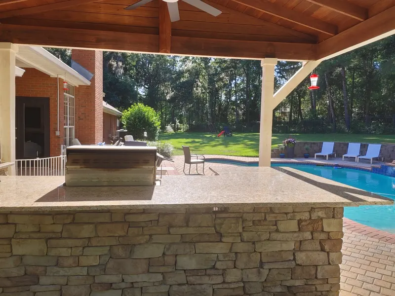 Light speckled stone counter on a stacked stone base under a covered patio, with a built-in grill station and a pool beyond