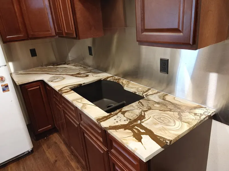 Cream counter with gold and brown swirls, a black sink and a metal backsplash above reddish cabinets